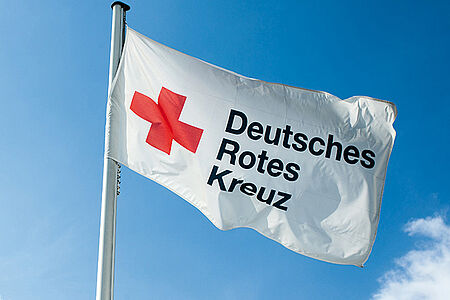 Eine Flagge weht im Wind vor einem blauen fast wolkenlosen Himmel. Darauf steht Deutsches Rotes Kreuz und ein rotes Kreuz ist zu sehen.