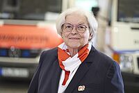 Rotkreuzlerin, förmliche gekleidet, vor einem Rettungsfahrzeug im leicht unscharfen Hintergrund