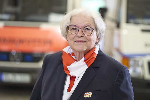 Helga Kuhlmann, förmliche gekleidet, vor einem Rettungsfahrzeug im leicht unscharfen Hintergrund
