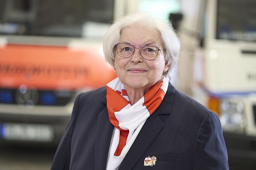 Helga Kuhlmann, förmliche gekleidet, vor einem Rettungsfahrzeug im leicht unscharfen Hintergrund