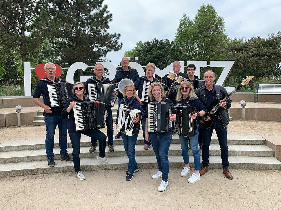 11 Personen stehen draußen in der Sonne auf einer kurzen Treppe vor dem Schild I Love Grömitz.